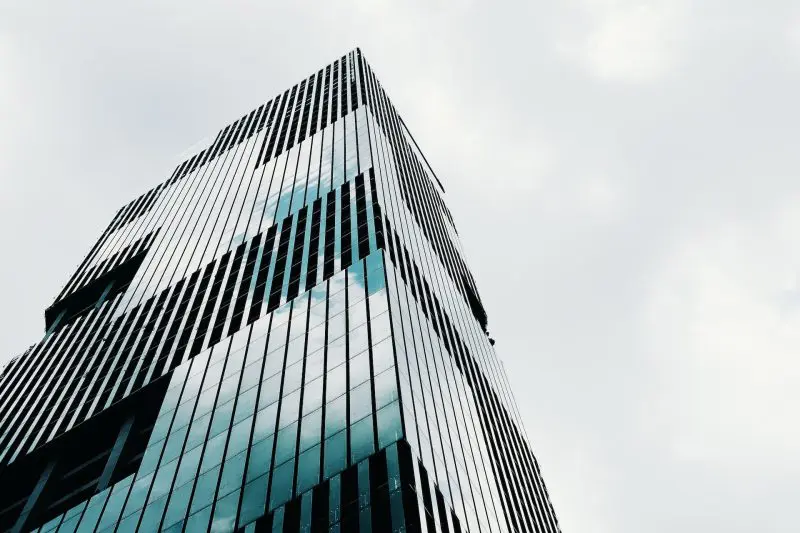 11-800×533.jpg Modern glass office building photographed from a low angle, showing reflective blue-tinted windows and vertical mullions against a pale sky.